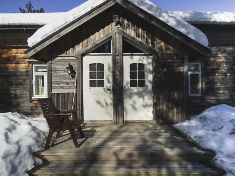 Eingangsbereich einer Holzhütte mit verschneitem Dach und Holzterrasse.