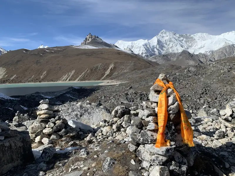 Gebetsfahnen und Steinmal in der Hochgebirgslandschaft mit See und schneebedeckten Bergen im Hintergrund.