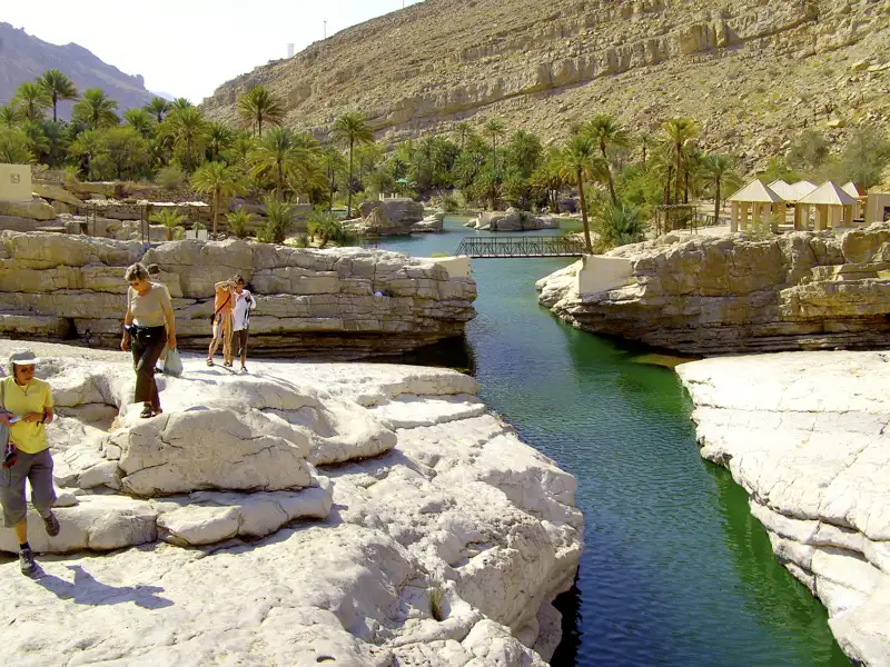 Reisende in einer Schlucht mit einem Flusslauf zwischen weißen Felsformationen und Palmen im Hintergrund.