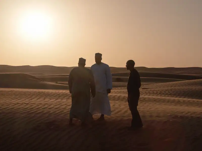 Drei Personen, zwei in traditioneller Kleidung, treffen sich in der Wüste bei Sonnenuntergang.