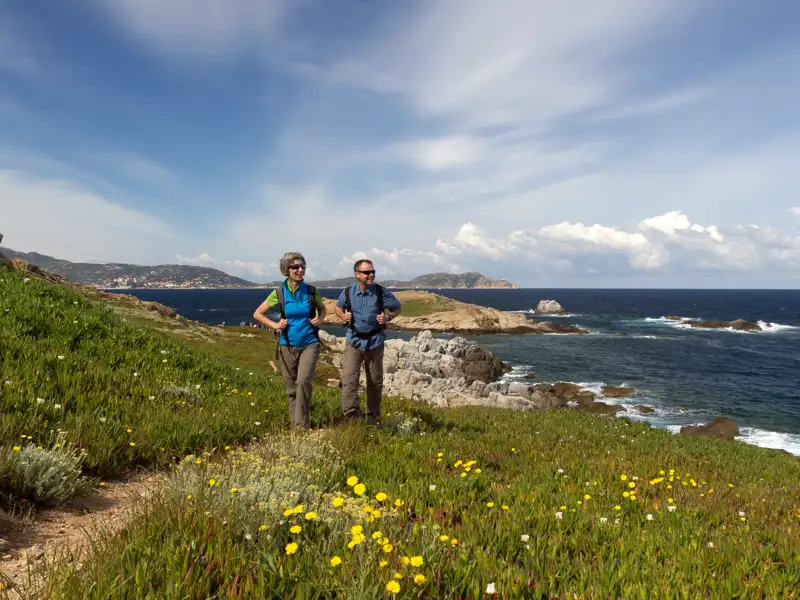 Zwei Wanderer auf einem blühenden Küstenpfad mit Blick auf das Meer.
