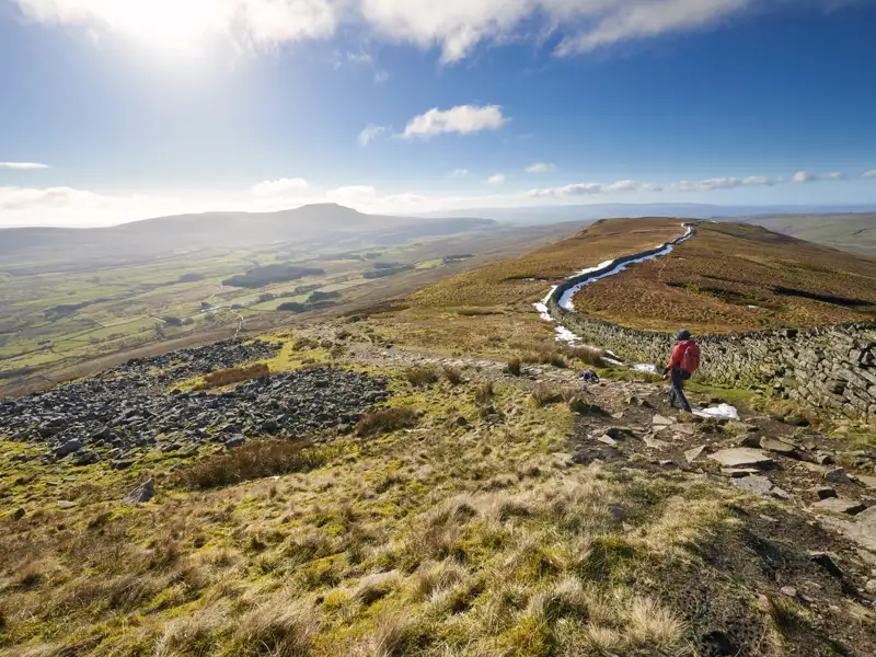 Wanderer auf einem Bergpfad mit Steinmauer und Talblick.