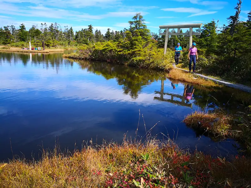 Zwei Wanderer passieren ein Torii am Ufer eines Bergsees.