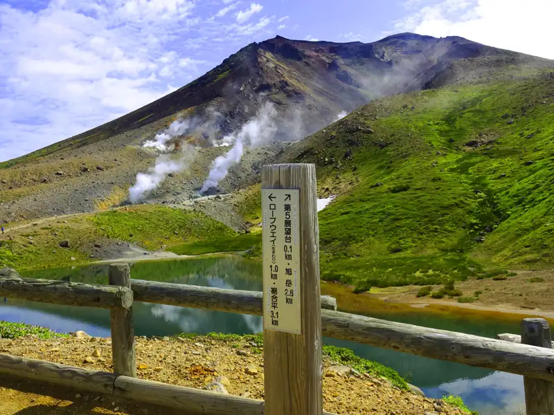 Wegweiser mit Entfernungsangaben zum fünften Aussichtspunkt, Asahidake und Seilbahn vor einem dampfenden Vulkan und einem See.