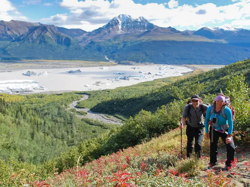 Zwei Wanderer mit Trekkingstöcken auf einem Bergpfad. Im Hintergrund ein Gletschersee mit treibenden Eisbergen und schneebedeckte Berge.