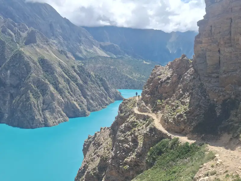 Wanderer auf einem schmalen Pfad entlang eines türkisfarbenen Bergsees, umgeben von steilen Felswänden.