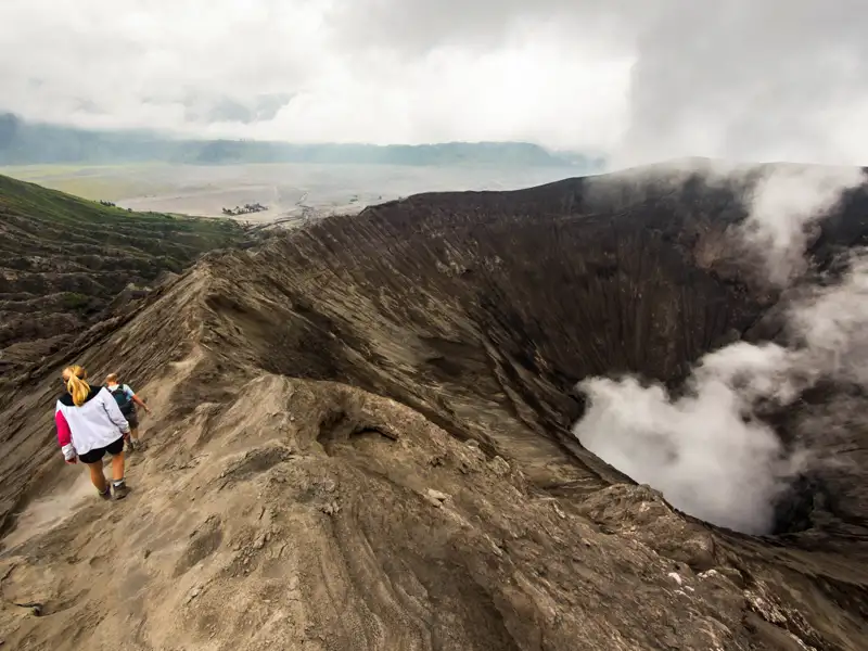 Wanderer am Kraterrand eines aktiven Vulkans mit Rauchentwicklung.