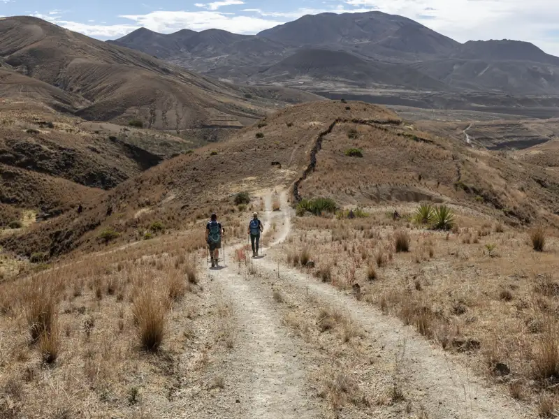 Zwei Wanderer auf einem Bergpfad in einer trockenen, bergigen Landschaft.