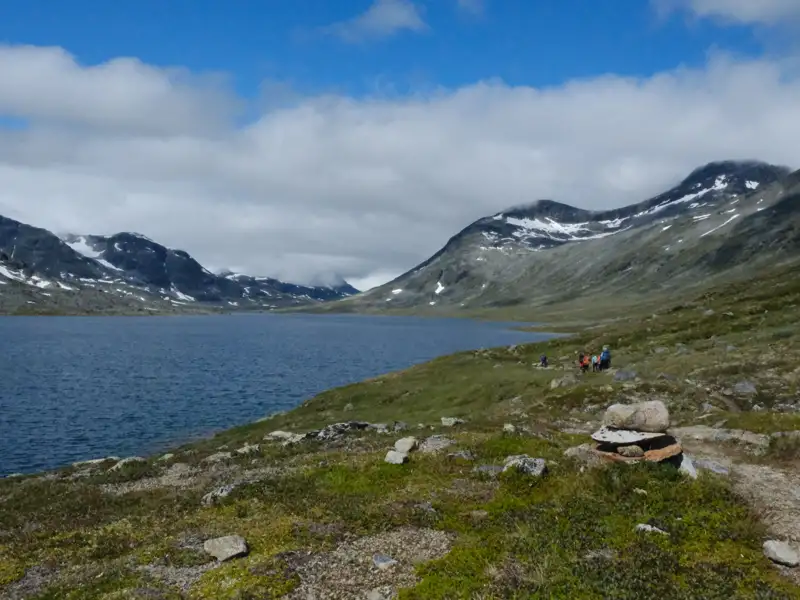 Wanderer auf einem Bergpfad neben einem See mit schneebedeckten Bergen im Hintergrund.