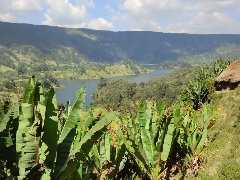 Panoramablick auf einen See inmitten von Hügeln mit Bananenstauden und einer Hütte.