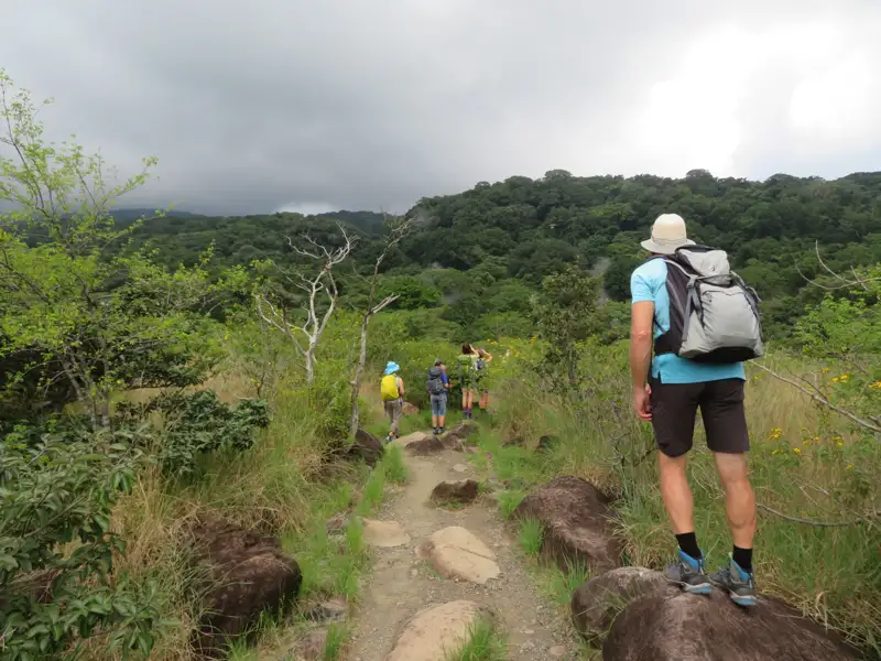 Wanderer auf einem Naturpfad, umgeben von Vegetation und mit Blick auf bewaldete Hügel.