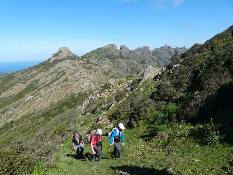 Drei Wanderer auf einem Bergpfad. Im Hintergrund sind felsige Gipfel und das Meer zu sehen.
