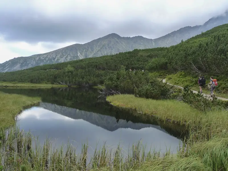 Zwei Wanderer auf einem Bergpfad neben einem See, umgeben von bewaldeten Hängen und Bergen.