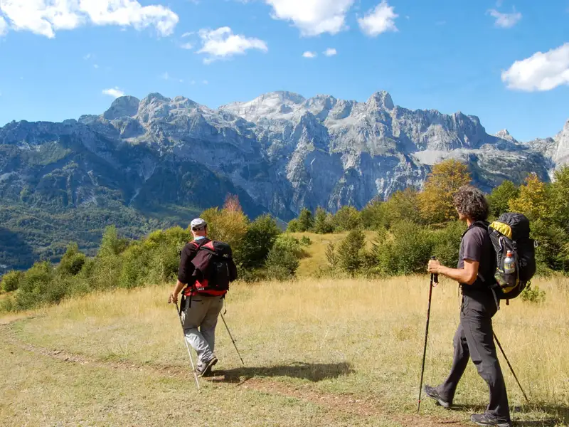 Zwei Wanderer mit Wanderstöcken auf einem Bergpfad mit einer Bergkette im Hintergrund.