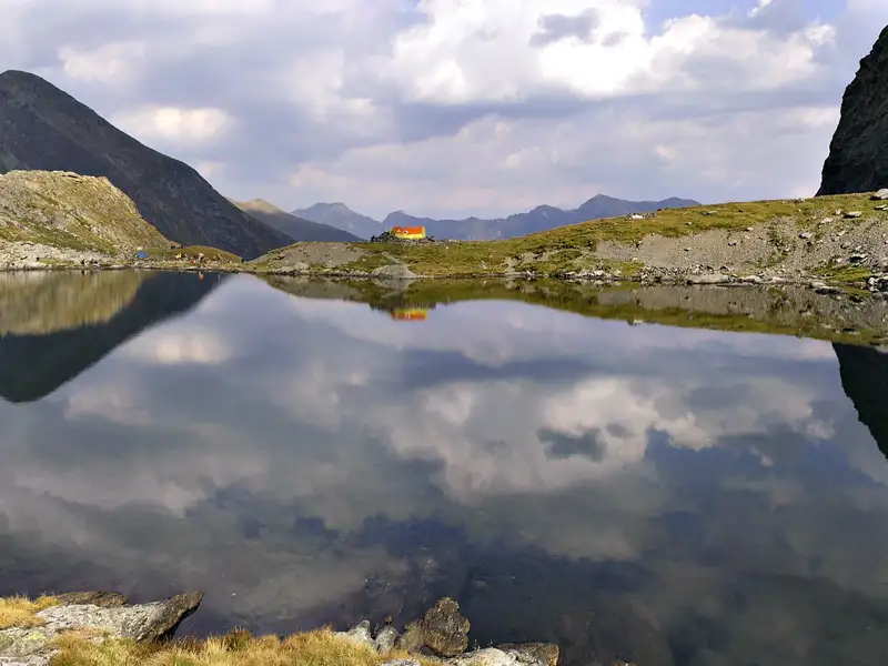 Idyllischer Bergsee mit Spiegelung der Wolken und einer Berghütte am Ufer.