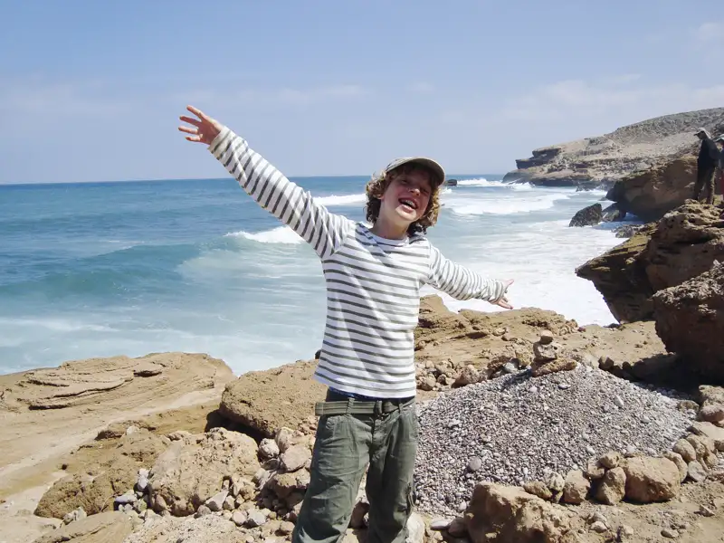 Kind mit ausgestreckten Armen an der felsigen Küste mit Blick auf das Meer.