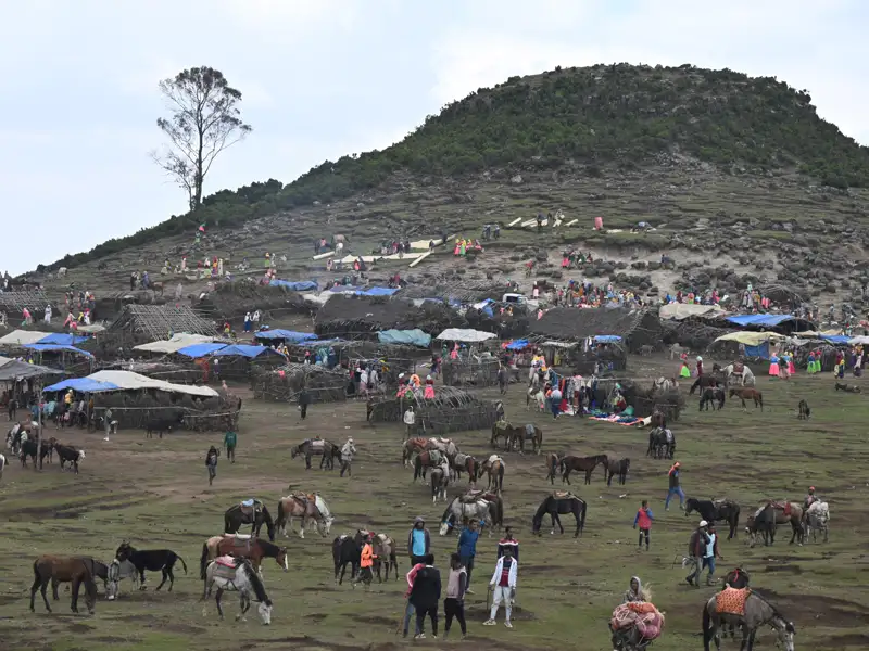 Ein belebter Markt mit Menschen und Pferden am Fuße eines Hügels, mit provisorischen Unterkünften und Zelten.