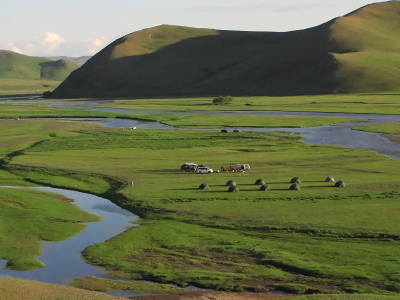 Blick auf ein Zeltlager inmitten einer grünen Flusslandschaft, umgeben von sanften Hügeln.