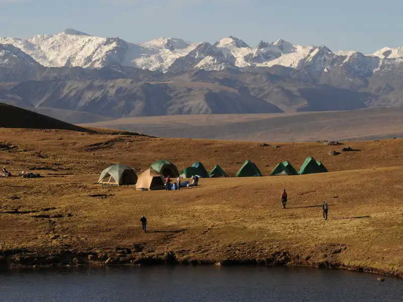 Wanderer zelten in einer hochgelegenen Landschaft mit einem See im Vordergrund und schneebedeckten Bergen im Hintergrund.