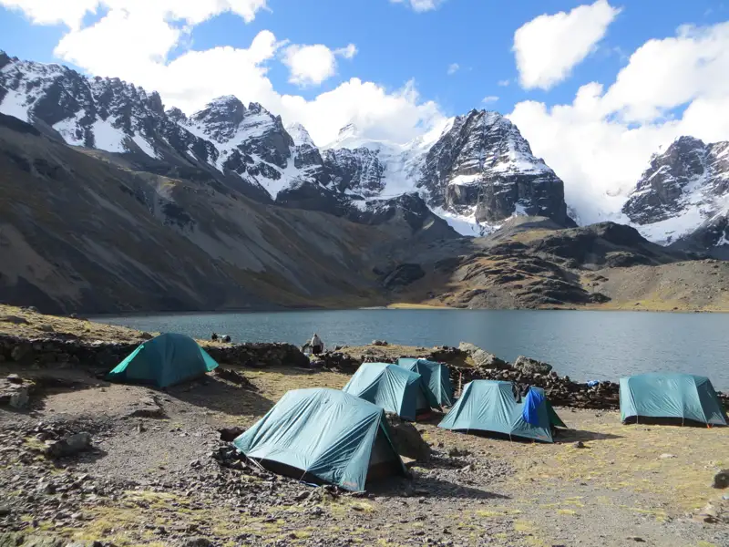 Zeltlager am Ufer eines Bergsees inmitten einer Hochgebirgslandschaft mit schneebedeckten Gipfeln.