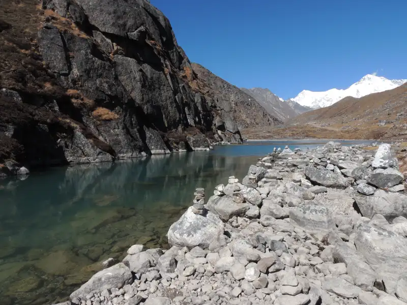 Klares Wasser eines Bergsees im Himalaya, umgeben von Felsen und mit schneebedeckten Gipfeln im Hintergrund.