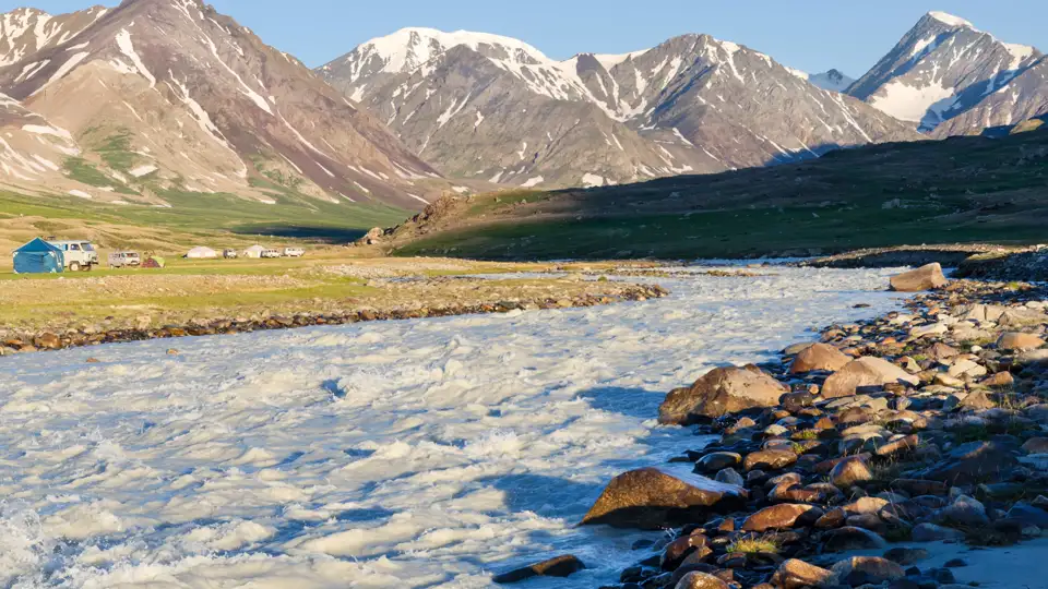 Schneebedeckte Berge, ein schnell fließender Fluss und ein Basislager im Vordergrund.